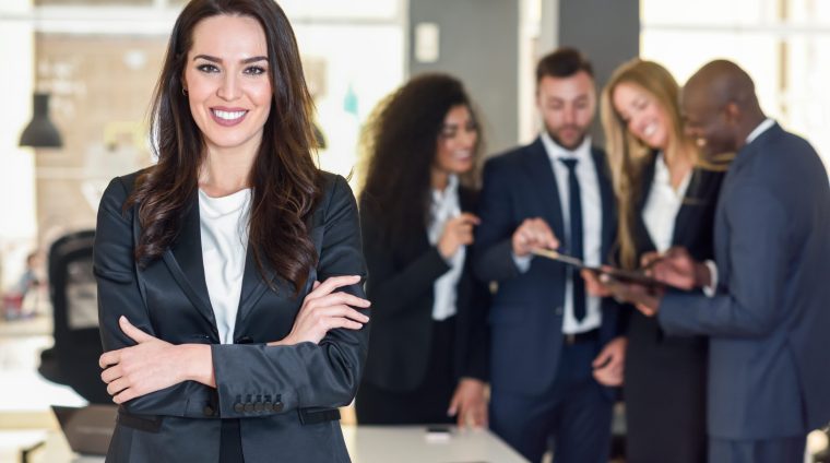 Businesswoman leader looking at camera in modern office with multi-ethnic businesspeople working at the background. Teamwork concept. Caucasian woman.
