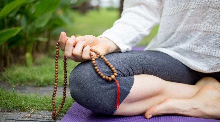 Close-up of hand of young woman wearing sweater and leggings sitting on mat, holding rosary beads and meditating