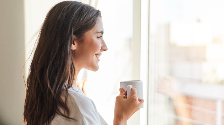 Portrait of a young woman drinking coffee when she has just woken up. She is looking out of the window of her house.
