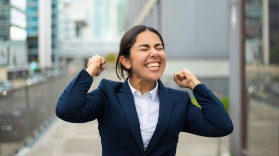 Excited businesswoman celebrating success. Cheerful young businesswoman in formal wear standing on street and raising fists. Winner concept