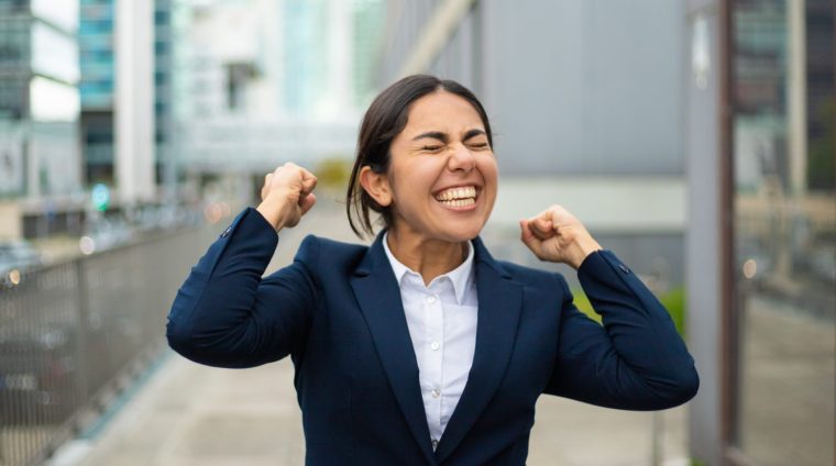 Excited businesswoman celebrating success. Cheerful young businesswoman in formal wear standing on street and raising fists. Winner concept