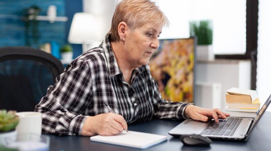 Mature entrepreneur taking notes on notebook working in home office. Elderly woman in home living room using moder technoloy laptop for communication sitting at desk indoors.