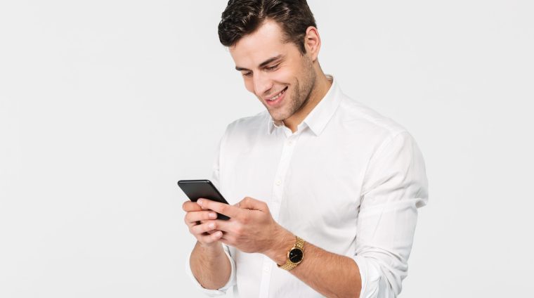 Portrait of a joyful smiling man in white shirt using mobile phone while standing isolated over white background